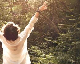 Person standing in a sunlit forest with arms raised, embracing a moment of joy and connection with nature.