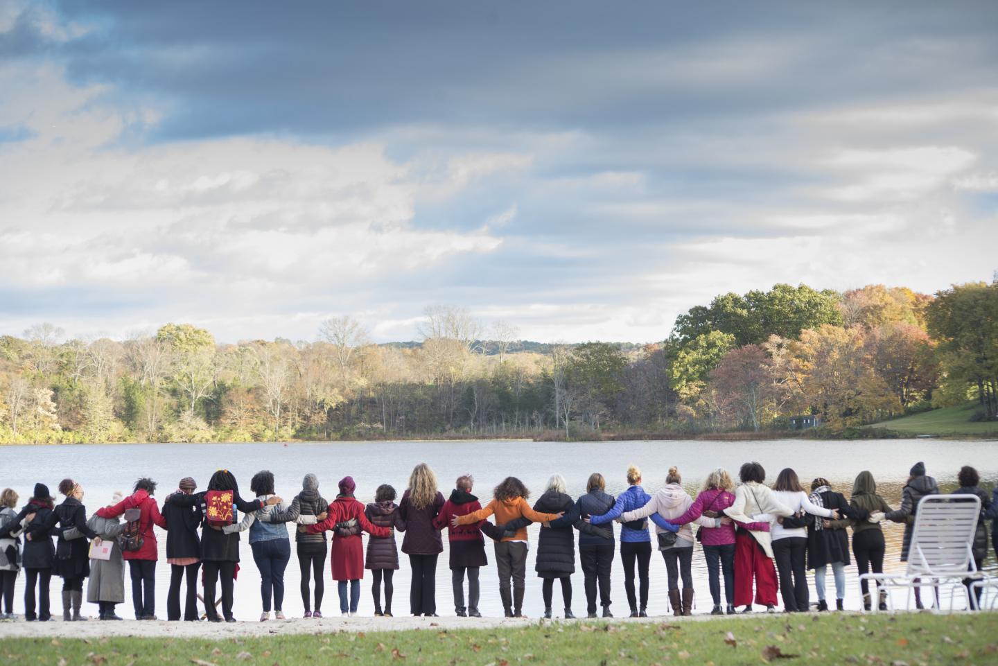 Many women standing arm in arm, looking over lake